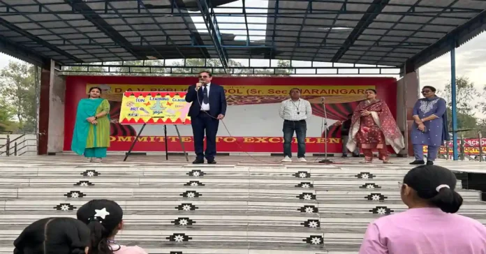 A school principal speaking on stage with teachers standing beside him during the launch of hobby classes, while students watch from the front.